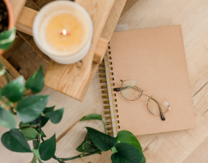 Photo showing a leafy vine plant, a blurry candle, and a pair of glasses on a beige notebook.